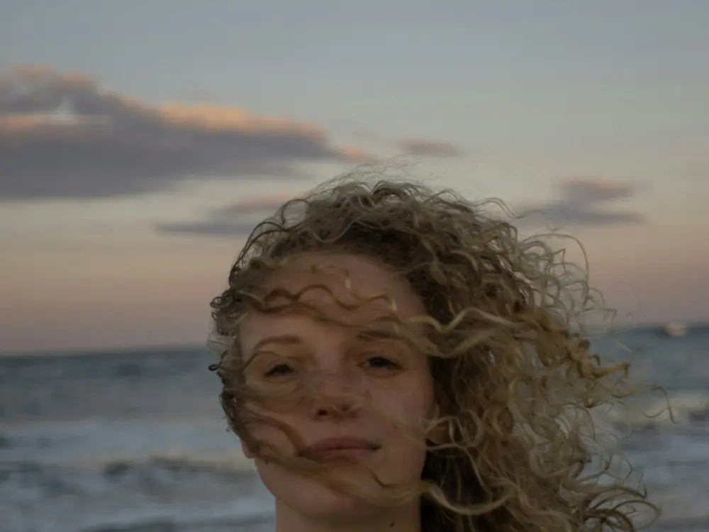 Woman with curly blonde hair windblown at the beach during sunset
